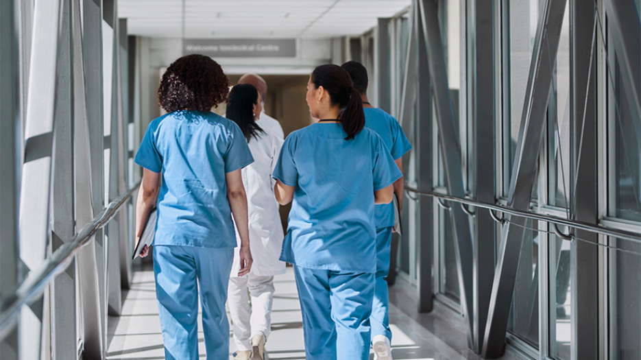 A group of nurses walking down a brightly lit hospital corridor photographed from behind.